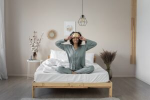 A woman wakes up in her bed looking happy and refreshed. She is wearing simple pyjamas, sitting up on the white sheeted bed with a wooden frame, and has her hands on a beige eye mask. Behind her is a minimalist decorated wall.